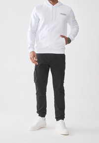 Mand iført hvid hættetrøje med forlomme og sorte cargobukser, stående med den ene hånd i lommen og hvide sneakers.