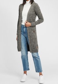 Grauer Strickcardigan mit langem Design, Knopfakzenten und zwei vorderen Taschen, kombiniert mit hellblauen Jeans und weißen Sneakern.