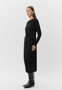 Kvinde med langt, glat hår iført en langærmet sort midikjole og sorte skinnende knælange støvler, stående foran en ensfarvet baggrund.