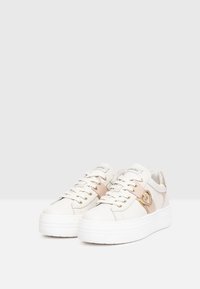 Baskets blanches avec une semelle épaisse blanche, des accents dorés et une texture lisse. Elles présentent des lacets et un détail de logo sur le côté.