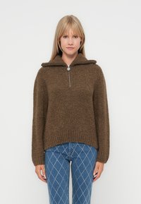 Brun strikket genser med glidelås og krage, med en teksturert overflate og senkede skuldre, kombinert med blå jeans med diamantmønster.