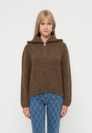 Pull en maille marron avec un col zippé, présentant une finition texturée et des épaules tombantes, associé à un jean bleu à motif diamants.