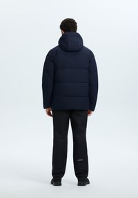 Marineblauwe gewatteerde jas met capuchon, voorzien van een gewatteerd ontwerp. Gepaard met zwarte broek en zwarte schoenen. Minimalistische stijl.