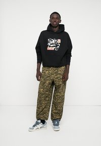 Svart hettegenser med grafisk trykk og frontlomme. Løse bukser i camouflagemønster med grønne nyanser. Joggesko med blå og hvite detaljer.