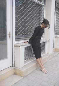 Maglietta nera a maniche lunghe con dettagli in pizzo, abbinata a pantaloni corti neri fino al ginocchio e sandali bianchi con fasce, su uno sfondo urbano.
