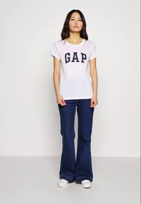 Kvinne med langt mørkt hår iført en hvit GAP t-skjorte, mørkeblå utvidede jeans og hvite joggesko, stående mot en nøytral bakgrunn.