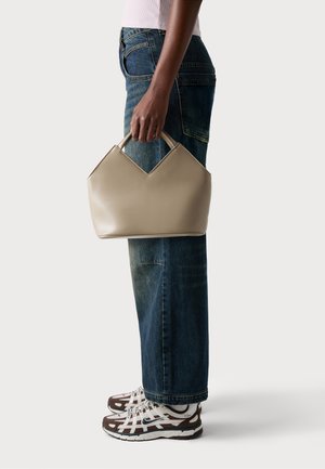 Beige leather handbag with a distinctive v-shaped top, matte finish, held by a person wearing blue jeans and patterned sneakers.