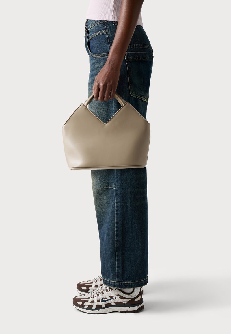 Beige leather handbag with a distinctive v-shaped top, matte finish, held by a person wearing blue jeans and patterned sneakers.