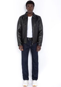 Schwarze Lederjacke mit Kragen, weißes T-Shirt darunter, dunkle Jeans und weiße Sneakers. Hände in den Taschen, neutraler Hintergrund.