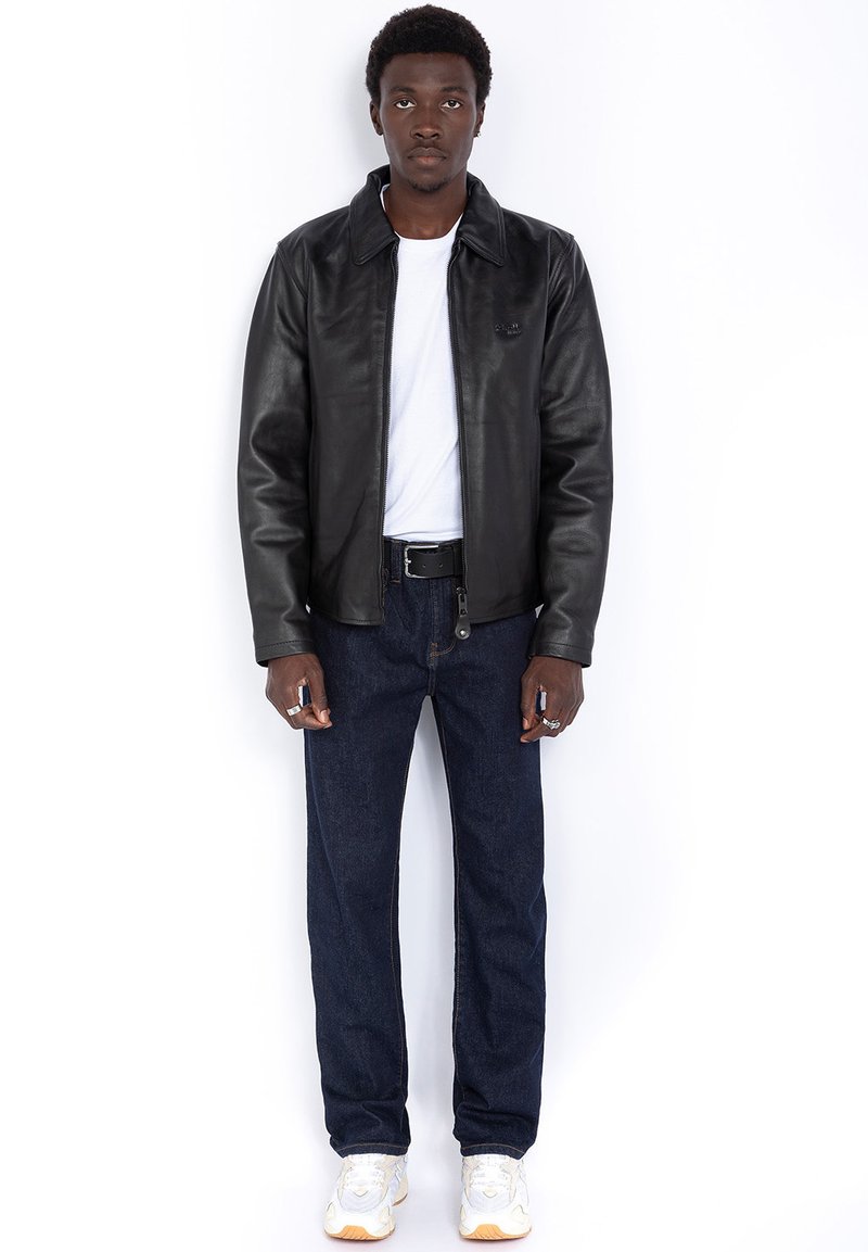 Schwarze Lederjacke mit Kragen, weißes T-Shirt darunter, dunkle Jeans und weiße Sneakers. Hände in den Taschen, neutraler Hintergrund.