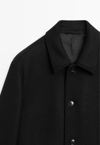 Zwarte wollen jas met een brede kraag, voorzien van een knoopsluiting aan de voorkant en een gladde textuur. Eenvoudig ontwerp, geen zichtbare patronen of accenten.