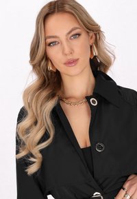 Musta trench-coat, jossa on lovittu kaulus, nappikiinnitys ja vyö. Mallilla on päällään kultaisia koruja ja hänellä on laineikkaat, olkapäälle ulottuvat hiukset.