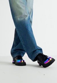 Sapatos pretos com sola grossa, apresentando um bico iridescente em arco-íris. Usados com jeans azuis largos, destacando o design do sapato.