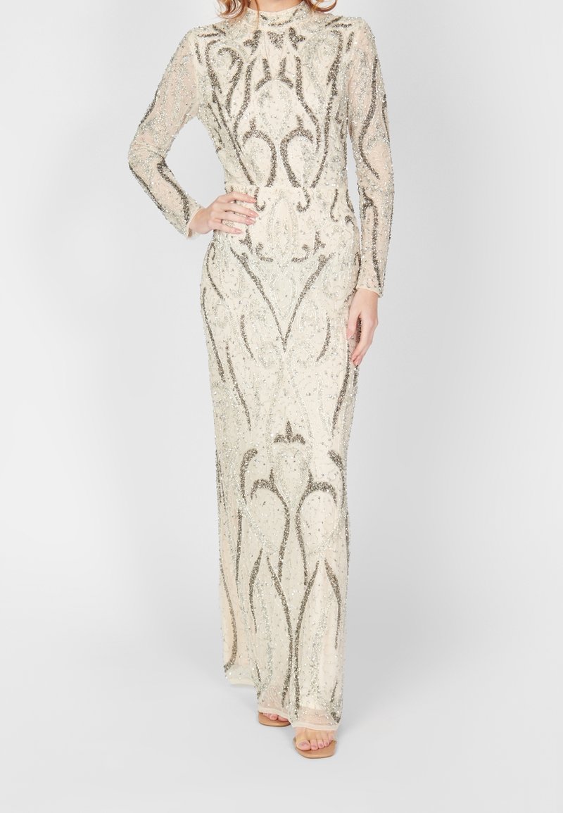 Femme portant une longue robe beige ajustée avec des motifs complexes de sequins argentés et des manches longues, debout devant un fond uni.