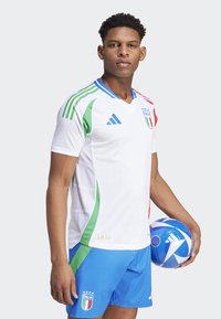 Biely futbalový dres so zelenými a červenými detailmi, modré šortky a logo talianskej vlajky; držiac modro-bielu futbalovú loptu s vzorom.