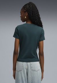 Camiseta ajustada de color verde oscuro con mangas cortas, escote redondo y textura suave, combinada con jeans de tiro alto de color azul claro.