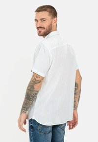 Hvid kortærmet skjorte med en krave, der kan knappes ned, fremstillet i et struktureret stof. Båret med blå jeans, der viser synlige tatoveringer på armen.