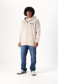 Beige zip-up hoodie met een voorzak en logo op de borst, gecombineerd met blauwe jeans en zwart-witte sneakers.