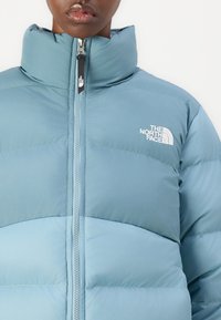 Šviesiai mėlynas pūkinis džiaisas su aukštu apykakle, užtrauktuku priekyje ir logotipu kairėje krūtinės pusėje; turi lygų paviršių ir siuvinėtą dizainą.