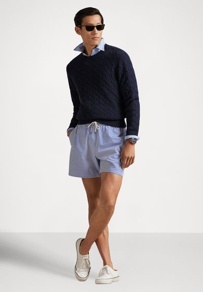 Tamsiai mėlynas megztinis su pynimais, šviesiai mėlyni marškiniai su apykakle, dryžuoti šortai, balti sportiniai bateliai ir saulės akiniai, demonstruojantys sluoksniuotą atsitiktinį vasaros stilių.