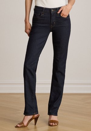 Kvinde iført mørkeblå jeans med lige ben, hvid top og brune hælede sandaler, stående på lyst trægulv.