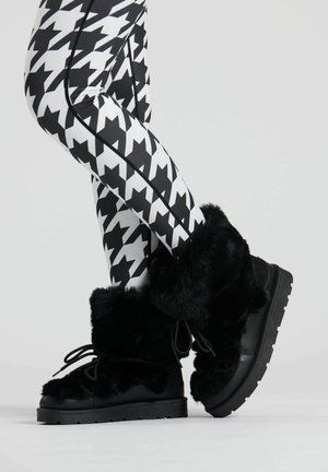 Black fur-lined boots with a textured upper, sturdy sole, and lace-up design, paired with high-contrast houndstooth patterned leggings.