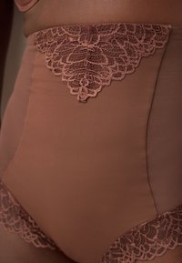Shorty gainant marron taille haute avec des détails en dentelle à l'avant, à la ceinture et aux ouvertures des jambes sur une personne debout.
