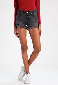 Pantalones cortos de mezclilla negra con dobladillos deshilachados, cinco bolsillos y costuras en contraste. Combinados con una camiseta de manga larga roja y zapatillas blancas.