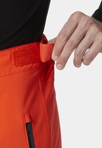 Rode buitenbroek met een klittenband taille-aanpassing. Voorzien van een zijzak met rits en een soepele stofstructuur. Handen die de taille aanpassen worden getoond.