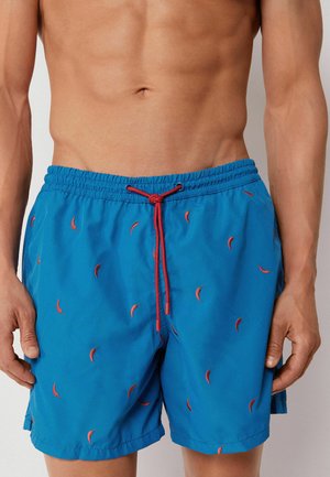 Shirtloser Mann trägt blaue Badeshorts mit roten Chili-Mustern und rotem Kordelzug in der Taille.