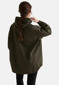 Persoon met gevlochten haar die een oversized donker groene hooded jas draagt en de capuchon aanpast, gezien van achter tegen een simpele achtergrond.