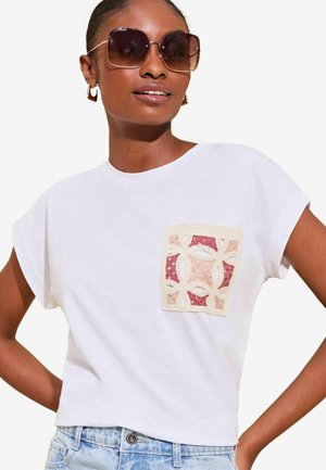 Kobieta w dużych kwadratowych brązowych okularach, złotych geometrycznych kolczykach, białym t-shircie z różowym i beżowym haftem oraz jasnoniebieskich dżinsach.