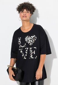 T-shirt noir oversize avec un design "LOVE" imprimé léopard, doté d'un col rond et de manches courtes. Le modèle tient un petit sac à main noir.