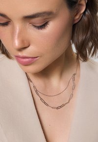 Warstwy naszyjników z wydłużonymi srebrnymi ogniwami, jeden grubszy i teksturowany, drugi cieńszy i delikatny, noszone na jasnobeżowym ubraniu.