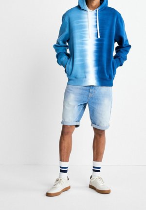 Mand iført blå og hvide tie-dye hættetrøje, lyse denim shorts med opkrogede kanter, hvide sokker med blå striber og hvide sneakers med brune såler.