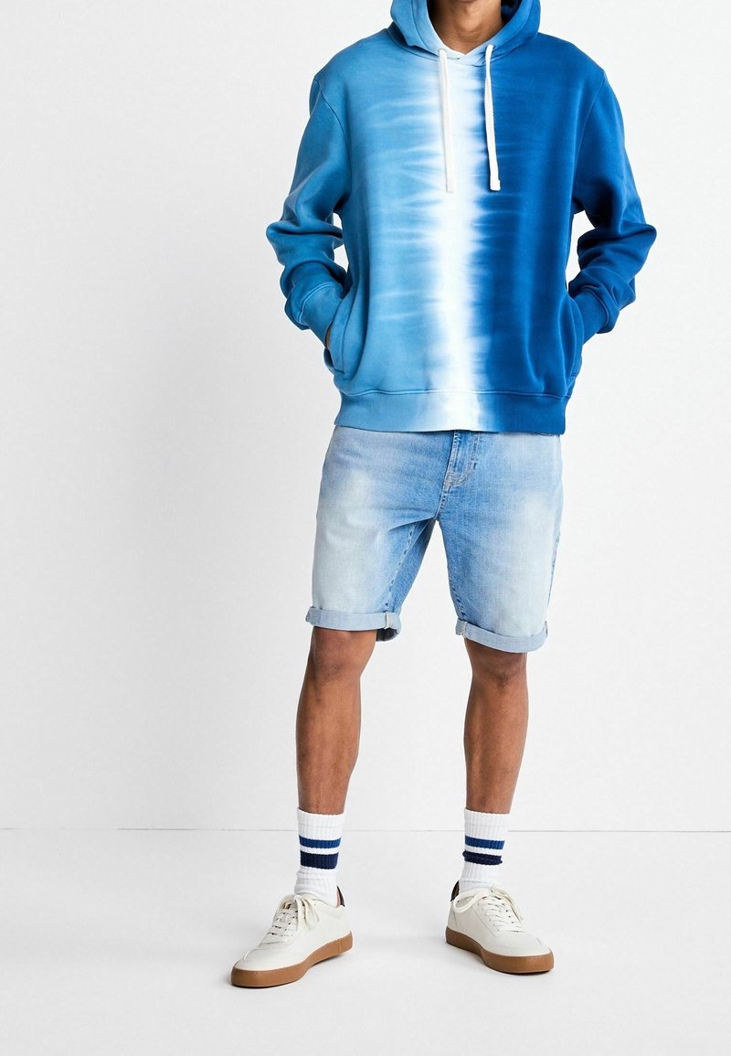 Homme portant un sweat à capuche tie-dye bleu et blanc, un short en denim clair retroussé, des chaussettes blanches à rayures bleues, et des baskets blanches avec des semelles marron.