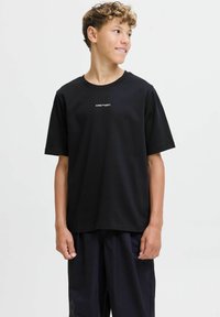 Černé bavlněné tričko s krátkým rukávem, kulatým výstřihem a bílým textovým logem na přední straně. Nosené s volně střiženými černými kalhotami.