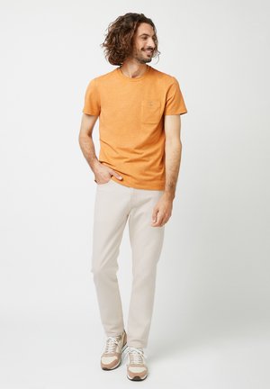 Man met krullend haar, gekleed in een oranje T-shirt met korte mouwen, lichtbeige broek en beige sneakers, staand met één hand in zijn zak en glimlachend.