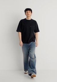 Νέος άνδρας που περπατάει, φοράει μαύρο υπερμεγέθες μπλουζάκι, φαρδιά μπλε τζιν και γκρι αθλητικά παπούτσια, σε λευκό απλό φόντο.