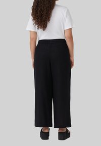 Schwarzweite Beinhosen mit einer lockeren Passform, aus einem leichten Stoff hergestellt. Mit einem elastischen Bund, der bis zu den Knöcheln reicht, kombiniert mit schwarzen Clogs.