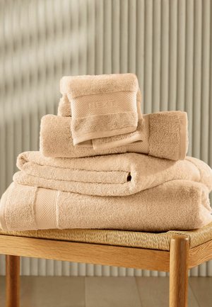 Beige foldede håndklæder stablet på et vævet sæde af en træskammel mod en stribet beige vægbaggrund.