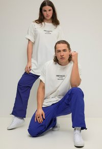 Twee personen in casual outfits. De ene draagt een wit T-shirt met zwarte tekst, en de andere draagt een blauwe broek en witte sneakers.