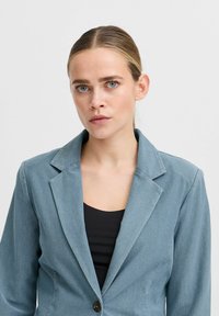 Svetle modrý blazer s výstřihy do špičky, na jednobodové zapínání a texturovanou látkou. V kombinaci s černým topem, který má jednoduchý design.