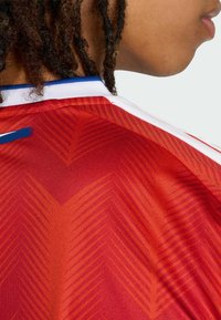 Zbliżenie na ramię i podbródek osoby ubranej w czerwony sportowy trykot z pomarańczowymi wzorami w kształcie szewronu oraz białym i niebieskim kołnierzem.