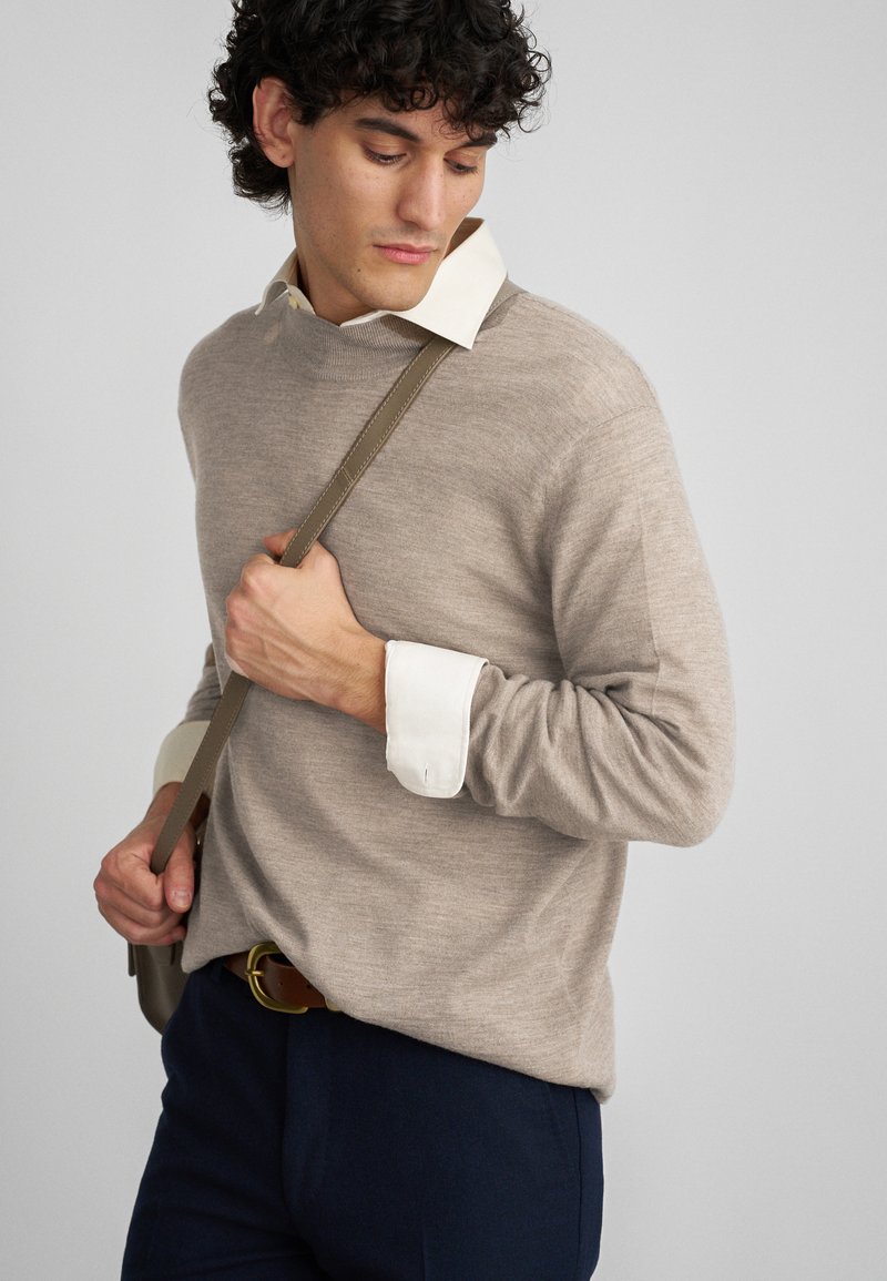 Bežs adīts džemperis ar apaļu kaklu, virsū uz baltas kreklu ar apkakli. Modelis tur bežīgu somu, ģērbies tumši zilās biksēs un ar brūnu jostu.