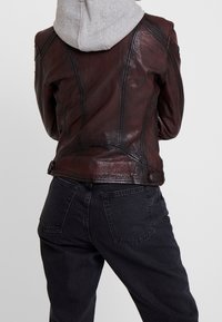 Person som bär en brun läderjacka över en grå hoodie, i kombination med svarta denimjeans, sedd från ryggen mot en enkel bakgrund.