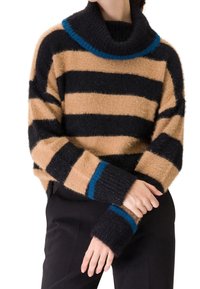 Personne portant un pull douillet et surdimensionné avec de larges rayures horizontales noires et beige, une bordure bleue au col et aux poignets, associé à un pantalon noir.