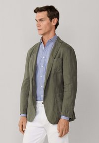 Mann trägt olivgrünen Blazer über blau-weiß gestreiftem Hemd mit weißer Hose, steht vor einfarbigem hellgrauem Hintergrund.