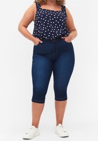 Top sin mangas de lunares azul marino con escote cuadrado y tirantes con volantes, combinado con pantalones capri de mezclilla azul oscuro. Zapatillas blancas visibles.