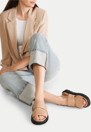 Béžové pantofle se silnou černou podrážkou, vybavené dvěma překrývajícími se koženými pásky. Nošeny s vykasanými světle modrými džínami a béžovým blazerem.